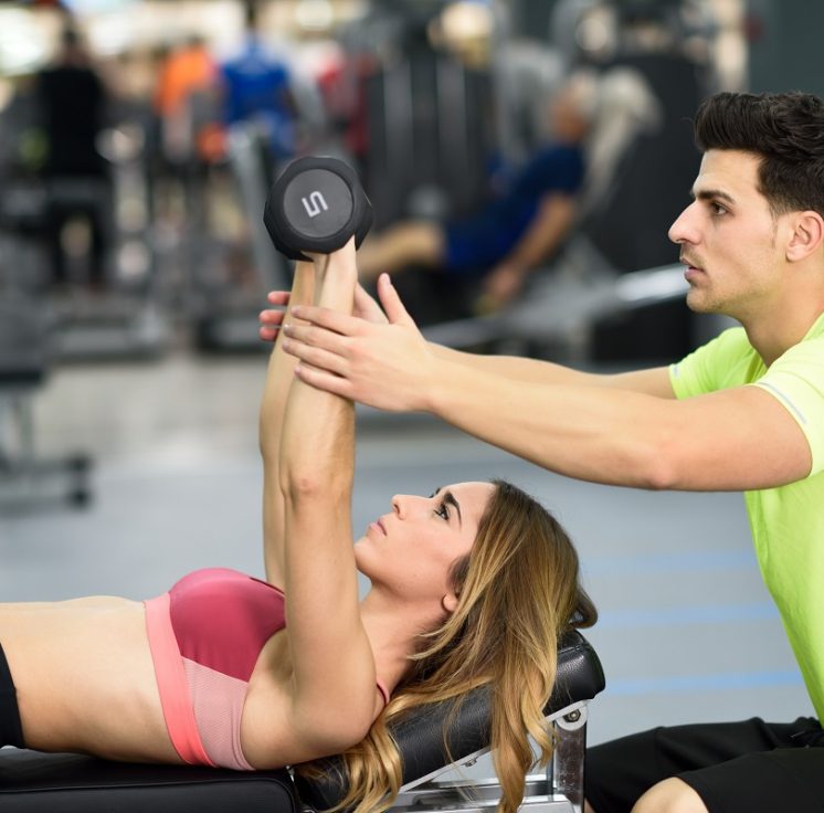 Personal trainer helping a young woman lift weights while working out in a gym